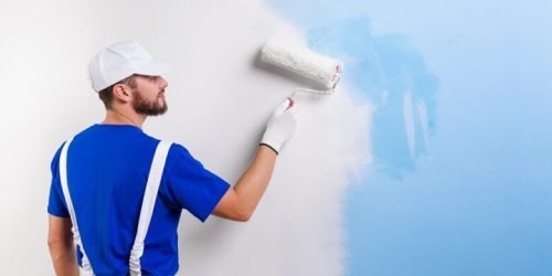 Back view of handsome young painter in white dungarees, blue t-shirt, cap and gloves painting a wall with paint roller.